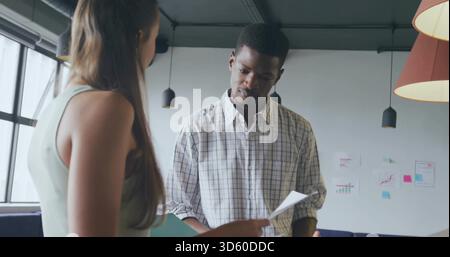 Reading male in checked shirt holding paper while female handing paper in office corner, charts Stock Photo