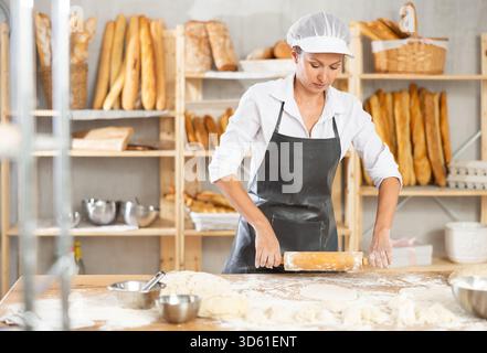 Making dough in bakery - female baker rolling out dough with rolling pin Stock Photo