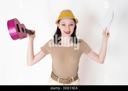Happy and smiling woman ukulele artist holding ukulele and sheet music in outstretched hands and looking at camera with positive facial expression. Fe Stock Photo