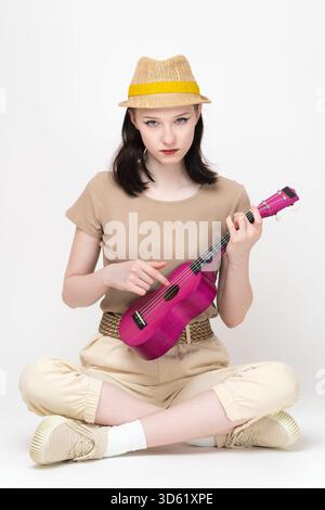 Young female musician holding ukulele and playing stringed musical instrument. Her gaze is fixed on camera. Woman ukulele player attired in casual clo Stock Photo