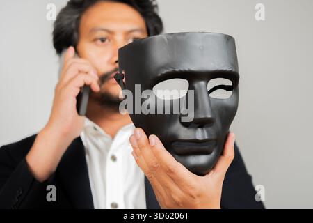 A business man using mobile phone while holding a black mask covering the insincerity of doing business together. Corruption concept. Stock Photo