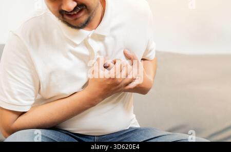 Cropped shot of adut man holding his chest and feeling pain suffering from heart attack while sitting on sofa at home. Stock Photo