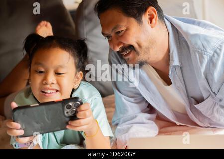 Father and daughters playing games together on the phone under the table at home. Happy family concept. Stock Photo