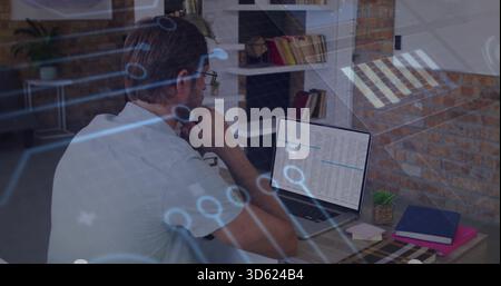Typing mid-adult man wearing white T-shirt and glasses at office desk, laptop showing data overlay Stock Photo