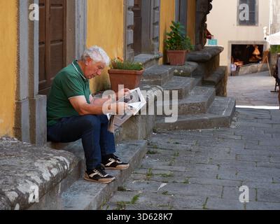 A man sitting on a door step reading the news paper in Castelnuovo Italy Stock Photo