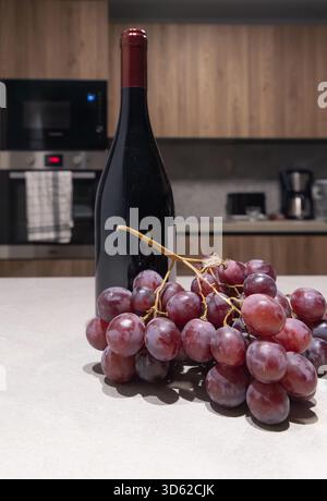 Close up vertical shot of a red wine bottle and grapes on a counter with a blurred, soft focus modern kitchen in the background Stock Photo