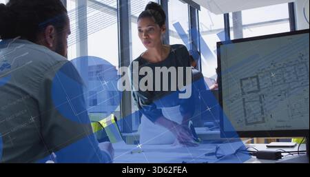 Pointing woman in dark top leaning over table in office, showing plans and screen with overlays Stock Photo