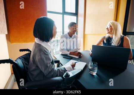 Three colleagues, one using a wheelchair, participate in a professional discussion around a table. This inclusive workspace celebrates diversity and f Stock Photo