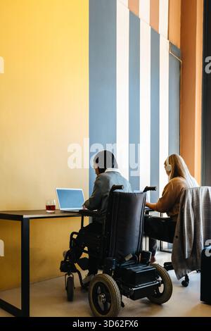 Two young women, one of them a wheelchair user, working on laptops in a modern office environment featuring colorful striped walls. The scene portrays Stock Photo