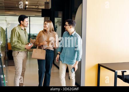 Three professionals discuss ideas while walking in a contemporary workspace. They hold devices and appear friendly, portraying teamwork, collaboration Stock Photo