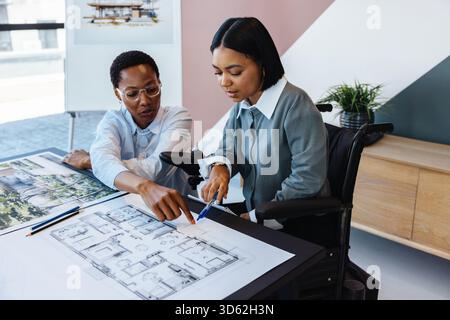 Two young women discussing architectural blueprints in a modern office. One, a wheelchair user, attentively collaborates with their colleague, showcas Stock Photo