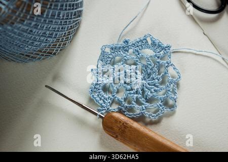 Close-up of a delicate light blue crochet snowflake motif in progress with a wooden-handled crochet hook and thread on a white background. Stock Photo