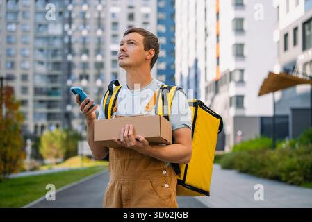 Young man courier wearing a delivery backpack and holding a brown parcel box while using a mobile phone, providing professional express shipping service in a modern urban area Stock Photo