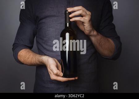 Man in grey-blue shirt holds dark red wine bottle with neutral blank label. Professional studio lighting, elegant black background. Well-groomed hands Stock Photo