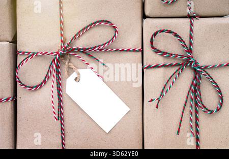 Background Stacked Christmas parcel boxes Close-up of white Tag for writing text on gift box wrapped in brown kraft paper tied with red, white and gre Stock Photo
