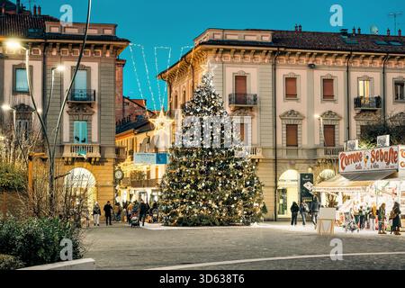 A brightly lit, large Christmas tree and a small holiday market stall stand in a cobblestone town square in Alba, Italy. Stock Photo