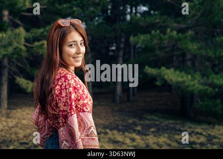 Young woman with long hair wearing a patterned shawl and sunglasses stands outdoors, smiling, in a natural forest environment. Stock Photo
