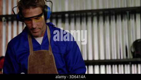 Inspecting welder working at bench in workshop, wearing coveralls, apron, safety glasses, earmuffs Stock Photo