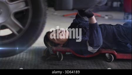 Lying female mechanic in dark coveralls inspecting under vehicle at garage, with red creeper, tire Stock Photo