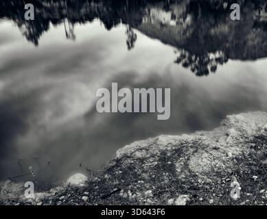 A dramatic, deep-toned black and white photograph focusing on the contrasting textures of the rugged shoreline in the foreground and the smooth, abstr Stock Photo