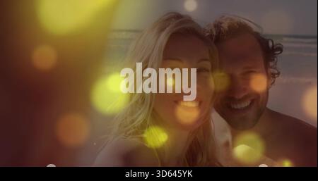Smiling woman wearing swimsuit leaning into man to her right on beach, holding smartphone, bokeh Stock Photo