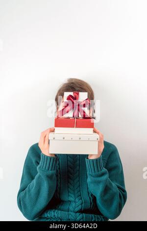 Girl in a teal knitted sweater holding a tall stack of red and white gift boxes with bows, fully covering her face against a white wall. Stock Photo