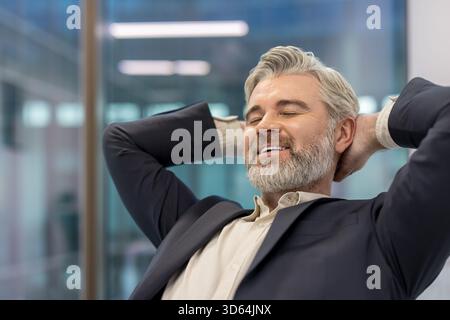 Mature businessman with gray hair and beard leaning back, eyes closed and smiling, feeling content and finding a moment of relaxation and satisfaction during a break at the office Stock Photo