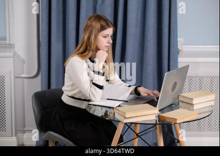 Woman sits at table surrounded by books, watching webinar on laptop, taking notes in notebook, working from home. Online student holds pen, jots down useful information in notebook. Remote work Stock Photo