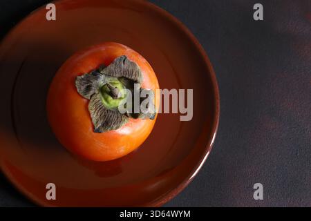 One ripe persimmon in a plate isolated on a brown background. Diospyros kaki. Healthy autumn fruit. Overhead view. Copy space. Stock Photo