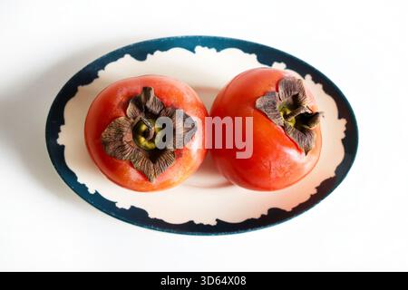 Two ripe persimmons in a plate isolated on a white background. Diospyros kaki. Healthy autumn fruit. Overhead view. Stock Photo