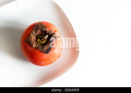 One ripe persimmon in a plate isolated on a white background. Diospyros kaki. Healthy autumn fruit. Overhead view. Stock Photo