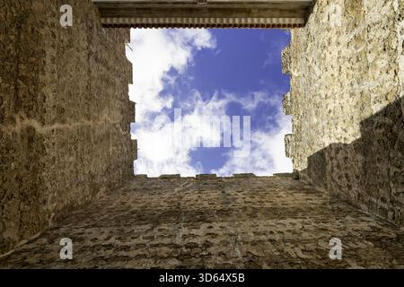 View of old stone walls reaching up to a square of bright blue sky with fluffy white clouds, creating a striking contrast in Óbidos, Leiria District, Stock Photo