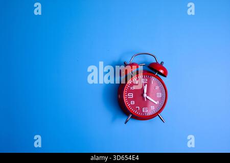 Red analog alarm clock on blue background highlights time awareness, urgency, and visual contrast in a minimalist composition. Stock Photo