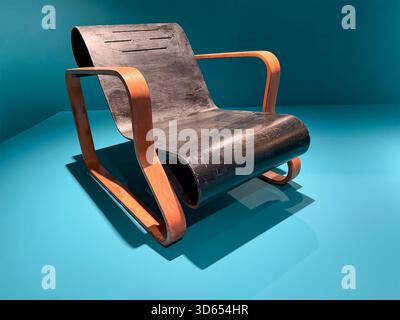 Vintage modernist armchair Paimio Chair by Alvar Aalto with curved plywood frame and black seat displayed against a turquoise background Stock Photo