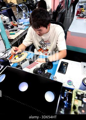Young asian technician working on electronic components and phones at a cluttered workbench in a tech repair workshop malaysia Stock Photo