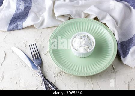 Homemade white sauce with herbs on an empty light green plate for your snacks. With a fork knife and napkin on the table Stock Photo