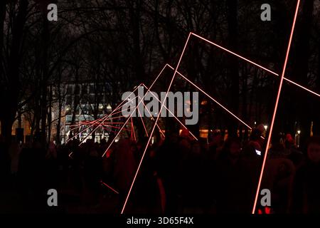 Nighttime outdoor installation featuring long angular red light lines stretching through park pathway with large crowd gathered underneath creating dy Stock Photo