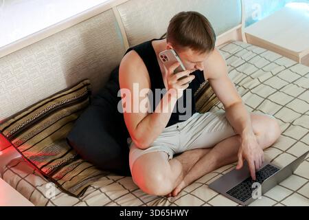 Relaxed young man sits on cozy bed, engaged in phone conversation while typing on silver laptop, soft lighting in modern bedroom Stock Photo