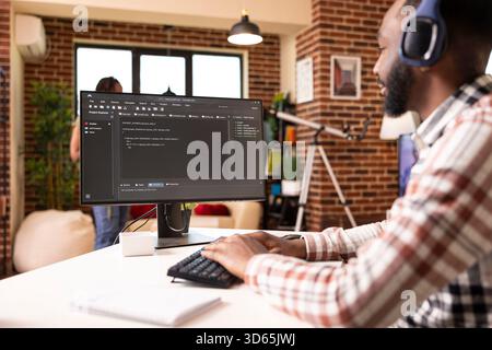 Remote male programmer sits at desk with headphones, coding and debugging scripts on computer. African american man listens to music and works on desktop pc, creating web application from home office. Stock Photo