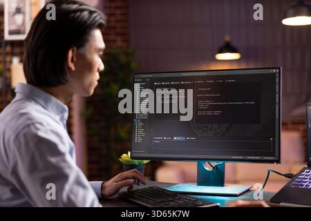 Self employed asian man in apartment home office focusing on coding scripts and software development using computers. Male programmer working remotely to build efficient websites and applications. Stock Photo