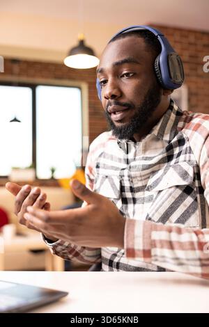 African American male freelancer with headphones gesturing during online video meeting from brick wall apartment. Self employed man using digital device, presents marketing project to remote clients. Stock Photo
