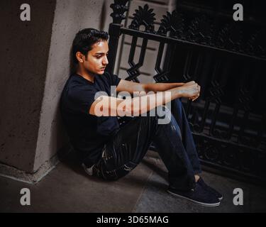 Contemplative Young Man Sitting Alone. A man dressed in casual jeans and a t-shirt sits on the floor, leaning against a wall. He gazes forward in cont Stock Photo