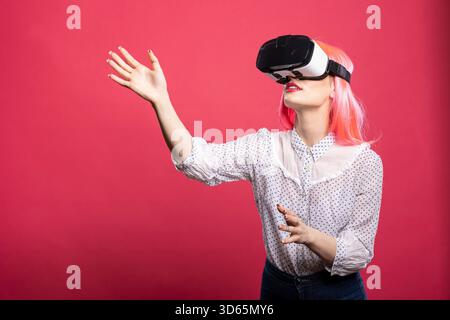 Young female gamer wearing virtual reality goggles, deeply engaged in high tech 3D simulation. Curious woman with VR headset, enjoying modern gaming through immersive digital technology. Stock Photo