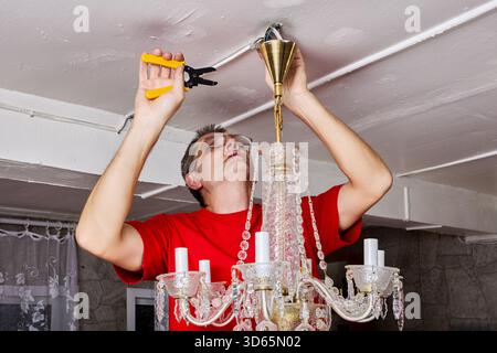 Electrician detaches old crystal chandelier, using pliers to disconnect wiring from ceiling fixture in rural house. Stock Photo
