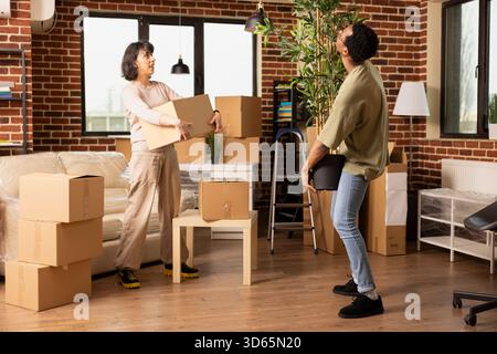 Caucasian woman holding brown box while watching african american boyfriend carry potted plant into new brick wall home. Multiethnic couple moving in together with packed belongings. Stock Photo