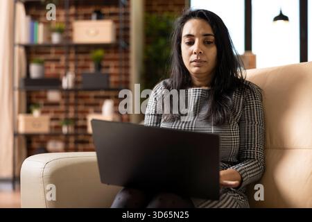 Indian businesswoman seated on cozy sofa, working remotely on laptop, reviewing financial reports and plans. Focused female freelancer using digital device, reading online articles in brick wall home. Stock Photo
