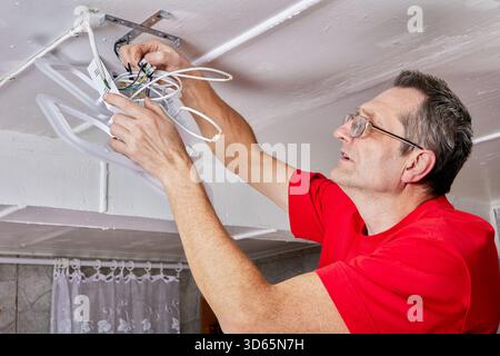 Worker installs LED chandelier on ceiling in countryside home, wires joined to contacts, mounting bracket secured, task nearing completion. Stock Photo