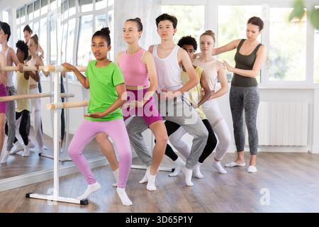 Dark-skinned girl practicing demi plie at barre with group Stock Photo