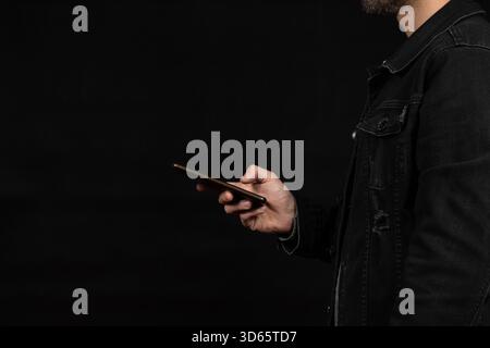 Close up to a man at the right side of the screen wearing a black denim jacket texting with a smartphone and a black background Stock Photo