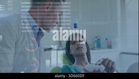 Explaining dentist in white lab coat leaning over patient in clinic chair, holding pink model HUD Stock Photo